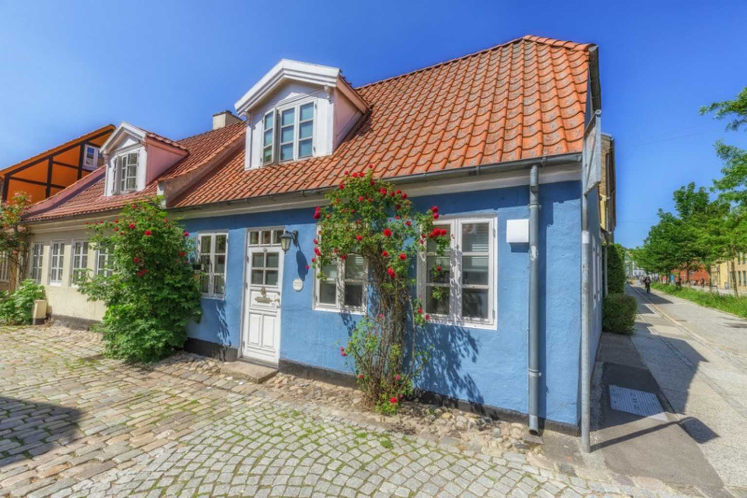 Lyseblåt byhus med rødt tegltag, kviste og røde roser ved facaden på en brostensbelagt gade