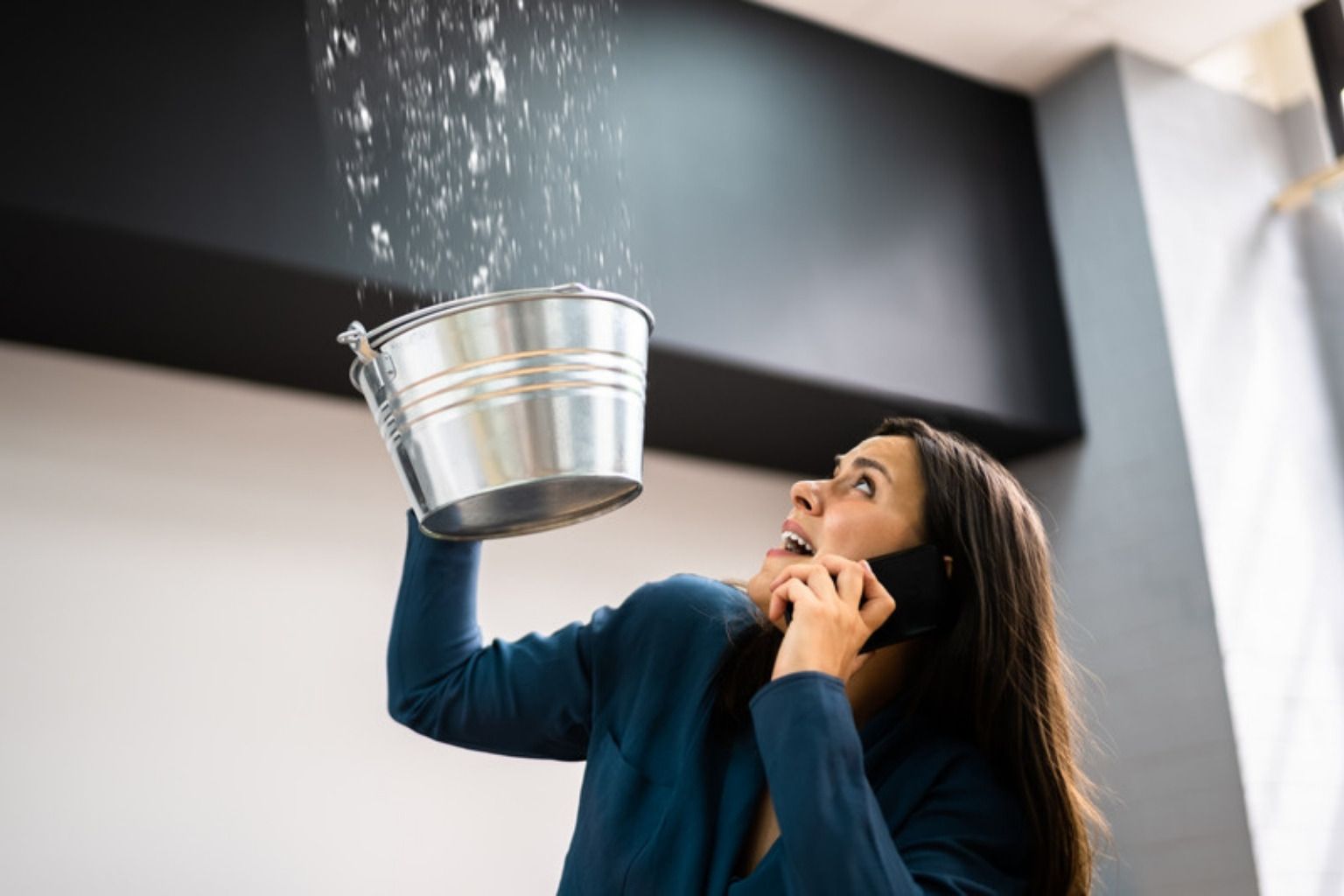 Kvinde holder en spand under vand, der drypper fra loftet, mens hun taler i telefon